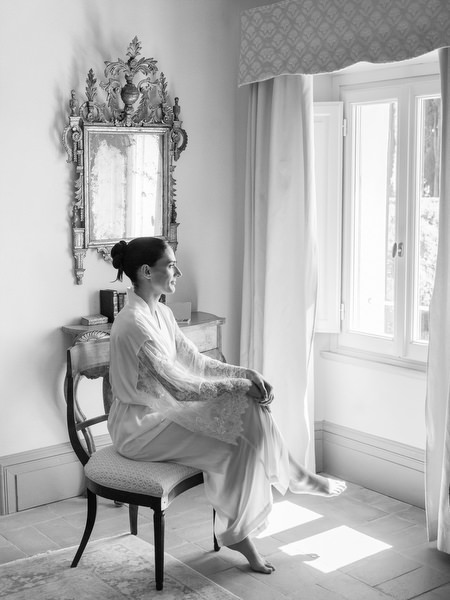 Bride in silk robe sitting by the window in soft morning light during bridal preparations in Tuscany