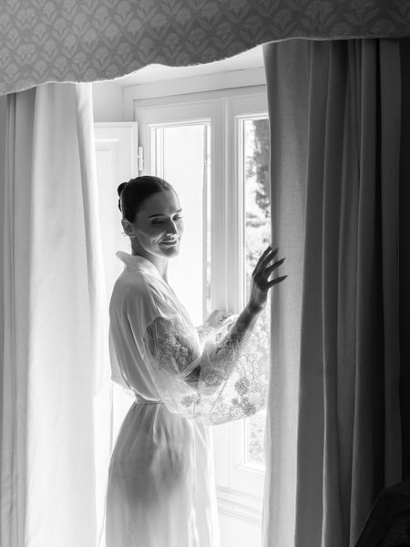 Bride in sheer robe opening curtains in natural window light during a luxury Tuscany wedding morning