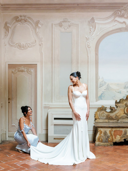 Bride in sculpted Galia Lahav gown as bridesmaid adjusts the train inside La Foce’s frescoed hall