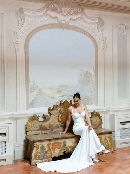 Bride in Galia Lahav gown seated on antique bench beneath frescoed archways at La Foce