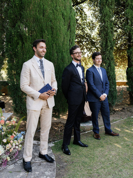 Groom and groomsmen standing beneath tall cypress trees before ceremony at La Foce estate