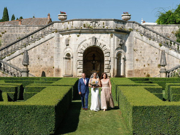 Bride walking down the hedge-lined aisle with her parents during luxury Tuscany wedding at La Foce