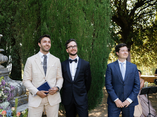 Groom and groomsmen standing beneath tall cypress trees before ceremony at La Foce estate