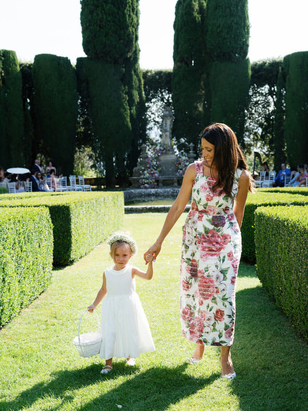 Flower girl holding basket while walking through hedge-lined aisle in Tuscan garden ceremony