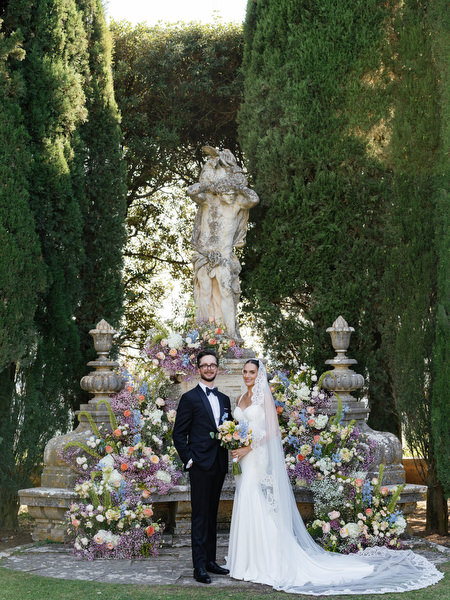 Bride and groom posing in front of abundant pastel floral installation at La Foce estate in Tuscany