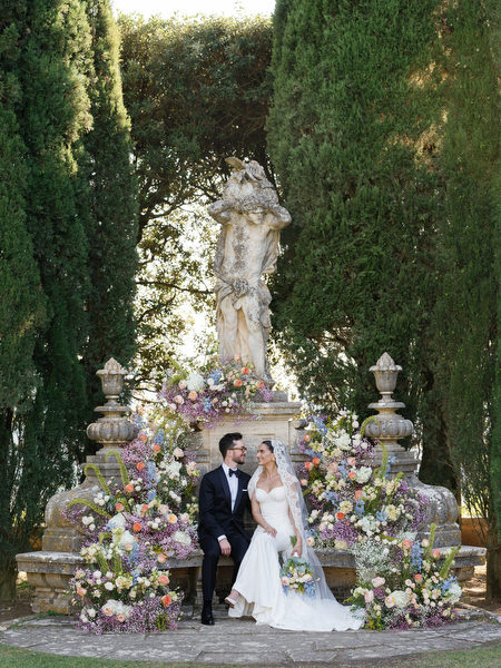 Bride and groom seated together beside classical statue and floral ceremony décor at La Foce