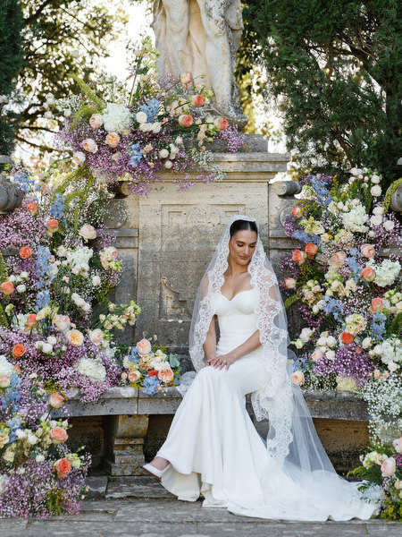 Bride in sculpted Galia Lahav gown seated beside abundant pastel florals at La Foce wedding in Tuscany