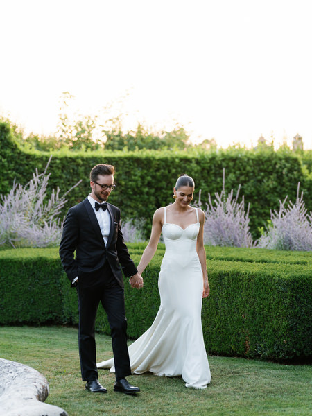 Bride and groom walking hand in hand through manicured gardens during golden hour in Tuscany