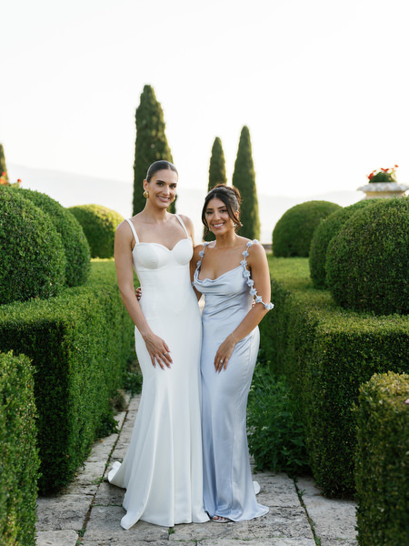 Bride and bridesmaid standing along hedge-lined garden pathway in Tuscany
