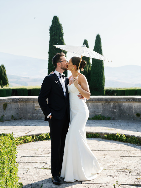 Bride and groom sharing a kiss beneath a parasol overlooking the Tuscan countryside