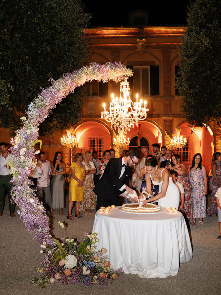 Bride and groom cutting wedding cake beneath crystal chandeliers at La Foce reception