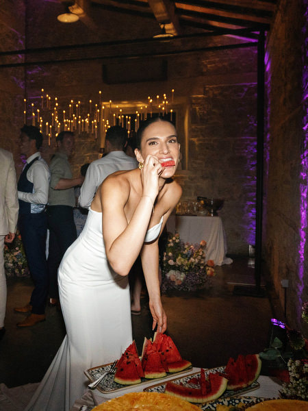 Bride laughing while holding watermelon slice during lively wedding after-party in Italy