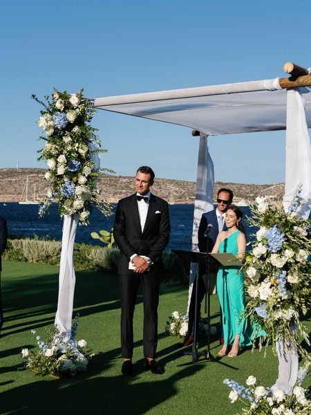 Groom standing beneath floral chuppah at Island Resort, ocean-view Athens Riviera wedding ceremony.