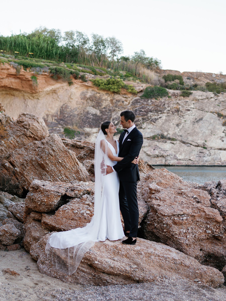 Bride and groom on rocky coastline at Island Resort, dramatic Athens Riviera seaside wedding portrait.
