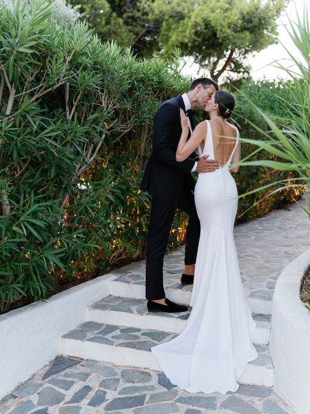 Groom kissing bride along garden path at Island Resort, romantic Athens Riviera wedding moment.