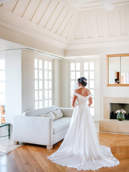 Bride in off-shoulder wedding gown standing in luxury suite at Anassa Hotel Cyprus.