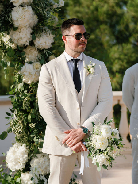 Groom standing beneath white floral arch at Anassa Hotel Cyprus wedding ceremony.