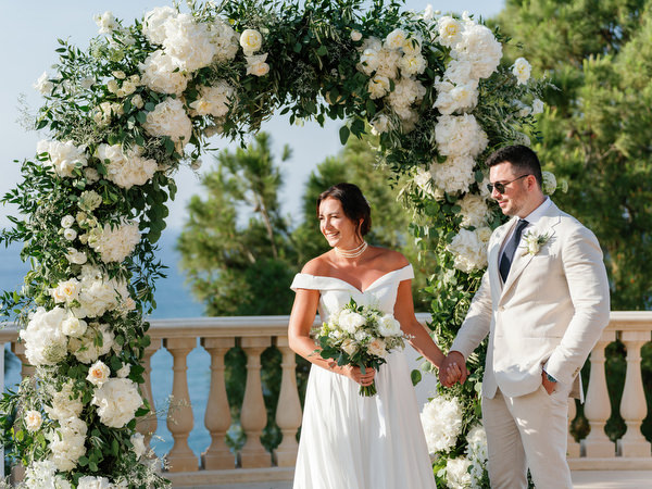 Bride and groom standing beneath white floral arch at Anassa Hotel Cyprus.