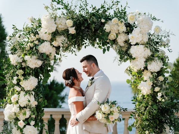 Bride and groom framed by white floral arch at Mediterranean resort wedding.