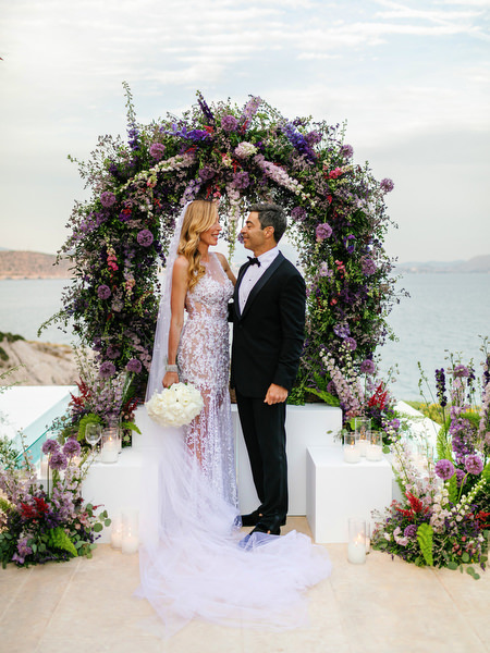 Newlyweds posing beneath a circular floral arch overlooking the sea at their Island Resort Athens Riviera wedding