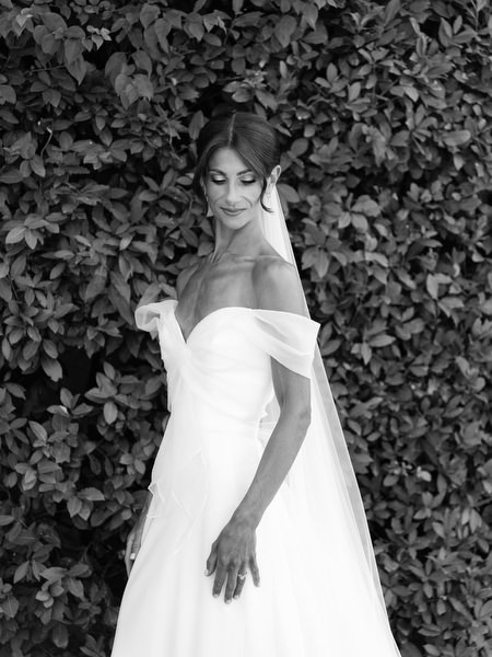 Bride in off-the-shoulder gown posing against greenery during luxury destination wedding in Greece