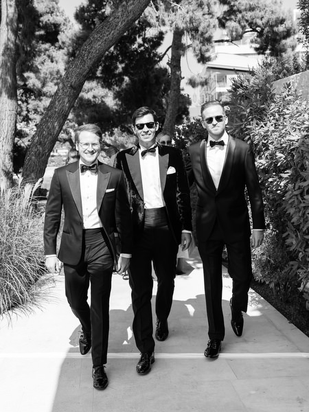 Groomsmen in classic black tuxedos walking through garden at Athenian Riviera wedding