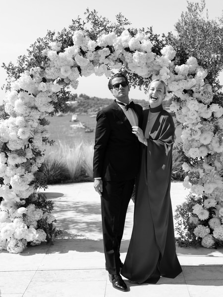 Black-tie wedding guests posing beneath white floral ceremony arch at Vouliagmeni Lake