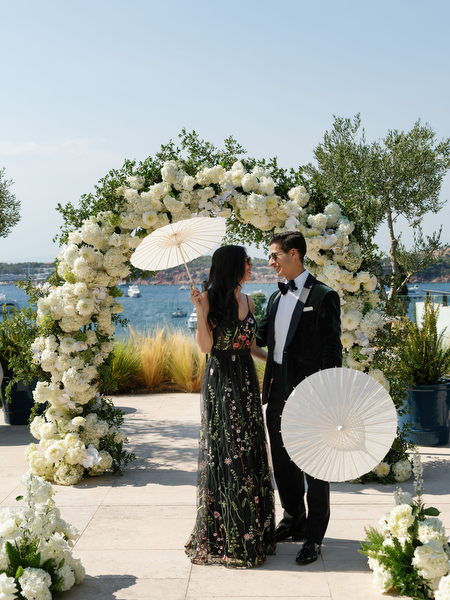 Wedding guests posing under white floral ceremony arch at Vouliagmeni Lake holding elegant parasols