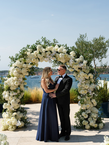 Black-tie wedding guests embracing beneath white floral arch overlooking Vouliagmeni Lake