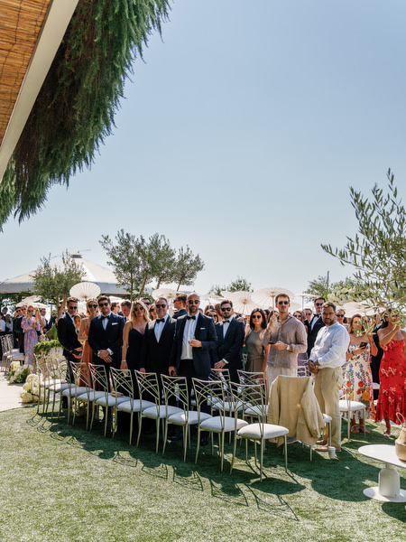 Wedding guests standing as ceremony begins at Vouliagmeni Lake overlooking the water