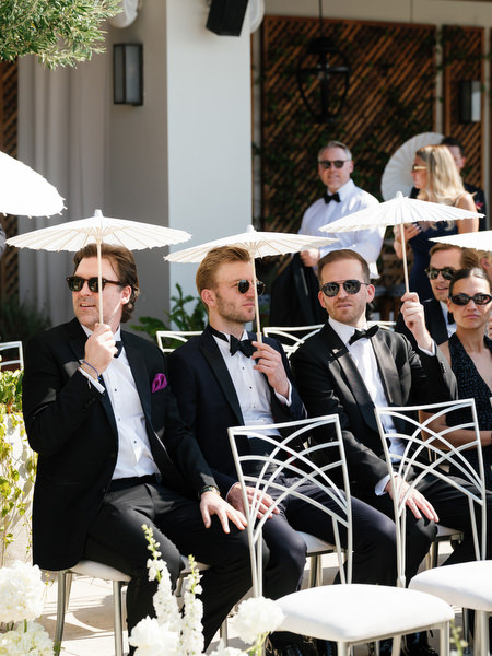 Black-tie guests seated in modern white chairs holding parasols during outdoor ceremony at Vouliagmeni Lake