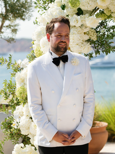 Groom in white dinner jacket standing before floral arch during black-tie Athens wedding