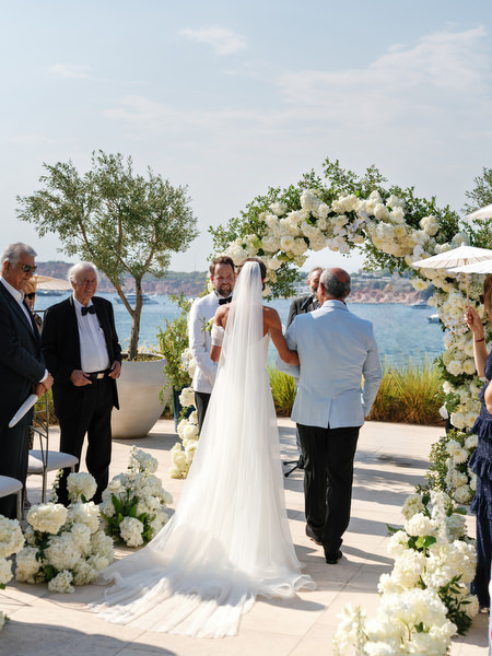Bride with long veil walking toward white floral arch at Vouliagmeni Lake ceremony