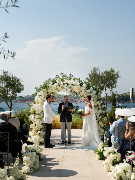 Bride and groom standing beneath white floral arch overlooking Vouliagmeni Lake during Athens wedding