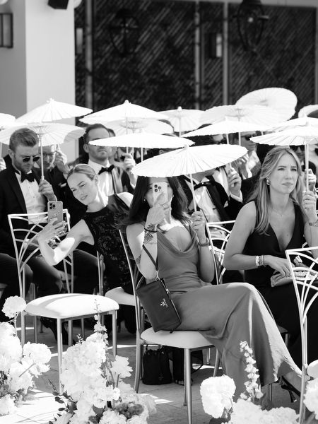 Black-tie wedding guests seated with white parasols during sunny outdoor ceremony at Vouliagmeni Lake