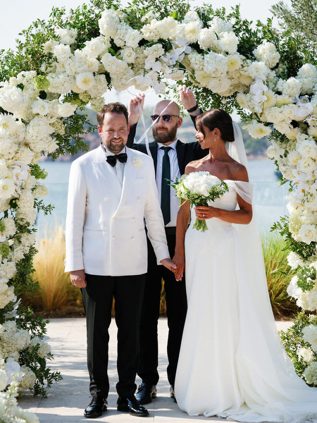 Bride and groom holding hands under circular white floral arch overlooking the water