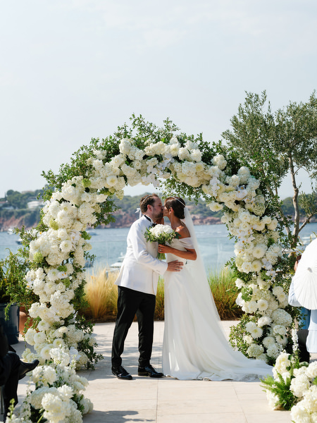 Bride and groom embracing beneath floral arch at Four Seasons Athens after saying “I do”