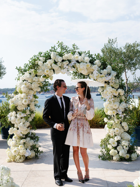Black-tie wedding guests posing beneath circular white floral arch at Four Seasons Athens