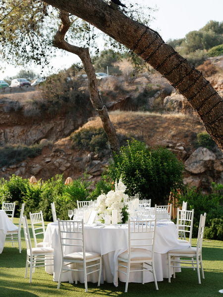 Round reception tables set with white florals beneath trees at Vouliagmeni Lake wedding