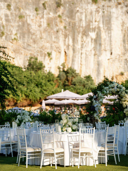 Elegant white reception tables arranged on lawn at Vouliagmeni Lake beneath dramatic cliffs