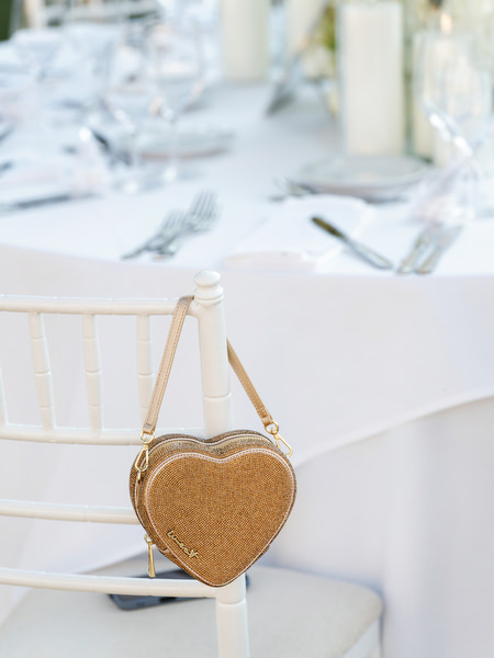 Gold heart-shaped evening bag hanging from reception chair at black-tie wedding