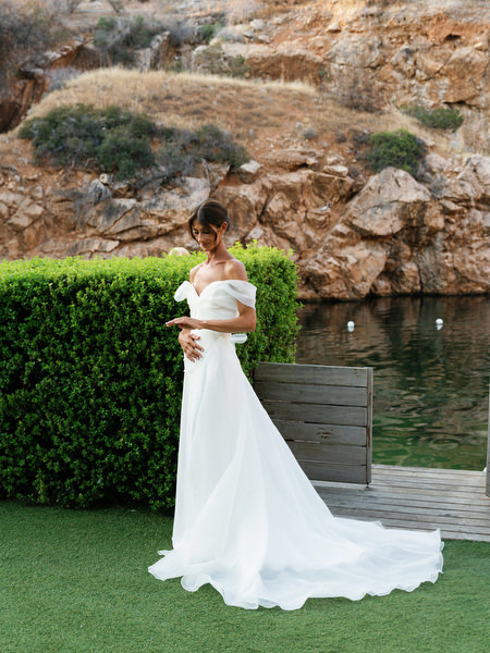 Bride holding bouquet beside manicured hedge and lake at golden hour