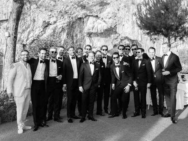 Large group of tuxedoed groomsmen posing together at lakeside wedding reception
