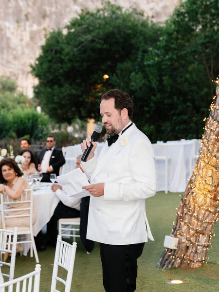 Groom in white dinner jacket delivering heartfelt speech at lakeside reception