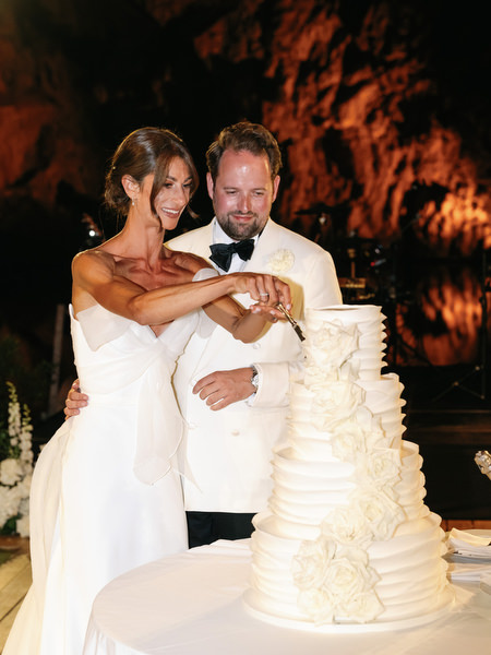 Bride and groom cutting multi-tier white wedding cake under warm evening lighting