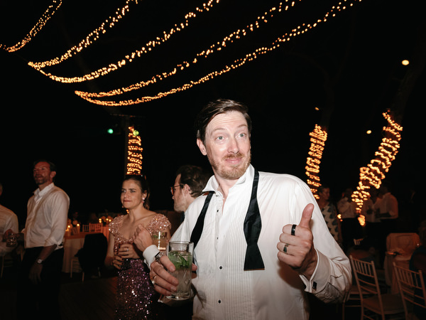 Smiling wedding guest giving thumbs up while holding drink on dance floor beneath glowing lights