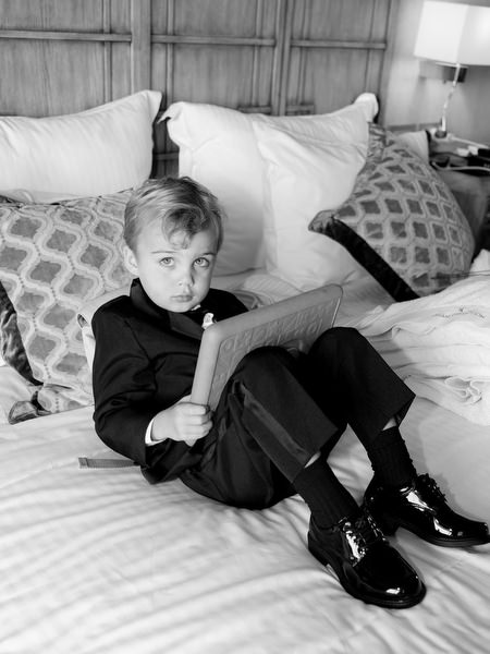 Young ring bearer sitting on hotel bed in black suit during Divani Apollon Palace and Thalasso wedding morning