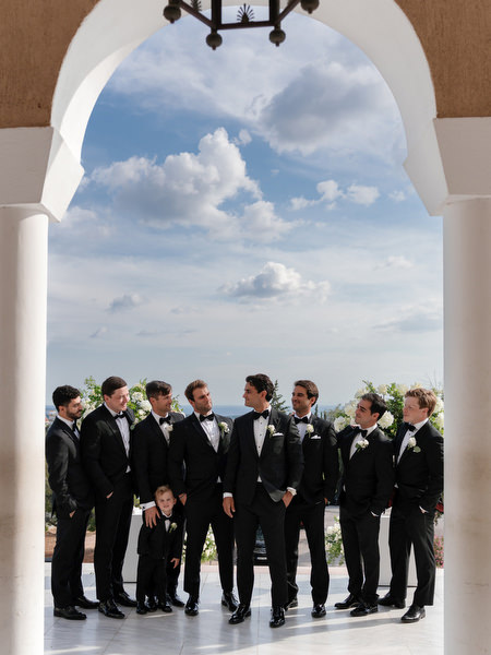 Groom and groomsmen in black tuxedos standing beneath church arch before Athens Orthodox wedding