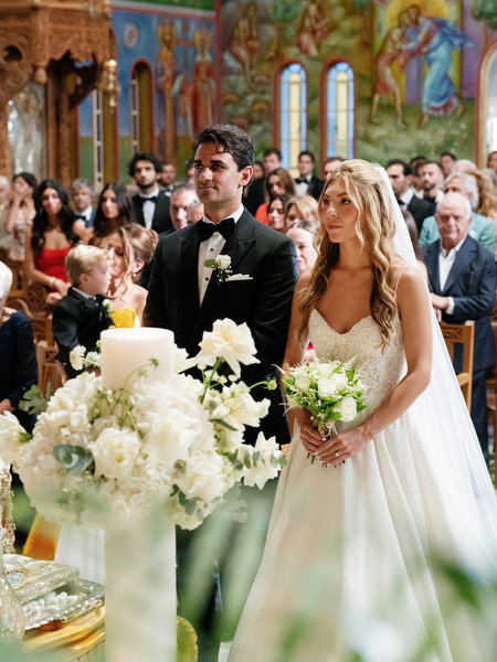 Bride and groom standing at altar surrounded by white florals during Greek Orthodox wedding in Athens