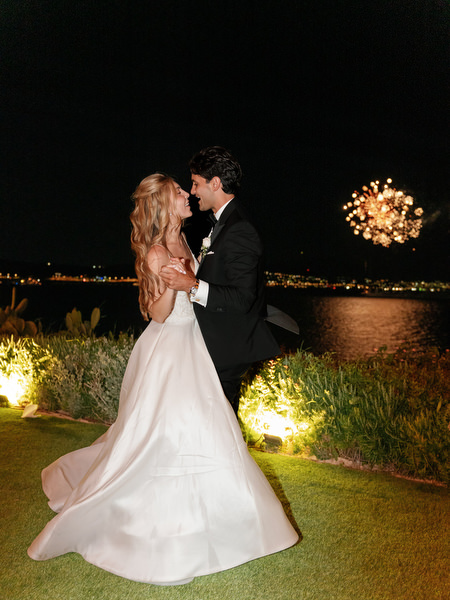 Bride and groom dancing outdoors at night with fireworks over the sea